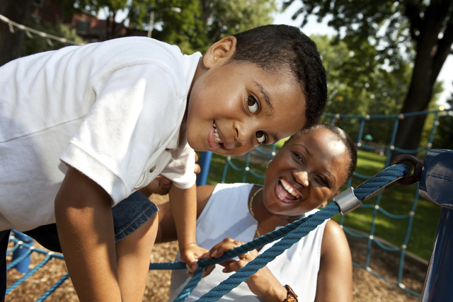 An African American boy climbs up a rope ladder on a playground as his mom smiles and looks on