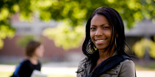 An African-American woman with long hair and a gray shirt stands outside and smiles at the camera