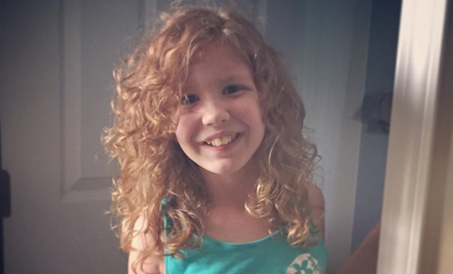 A girl with shoulder length curly red hair smiles at the camera in her home