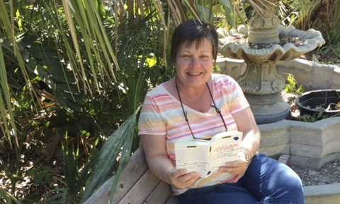 Debbie Rudy-Lack smiles as she sits on a bench holding up an open book