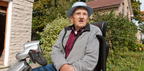 An elderly white man wearing a light blue hat sits on a mobility scooter outside a house