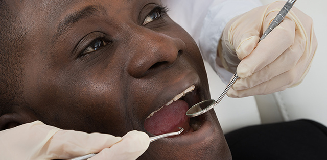 A dentist's hands hold tools in a Black man's mouth as he sits in a dentist's chair