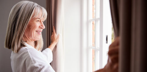 An older woman smiles as she stands at an open window