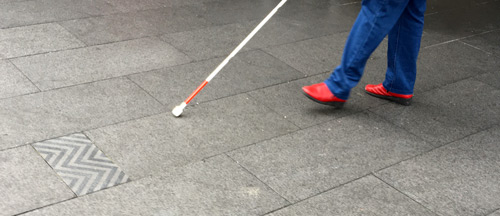 A person wearing jeans and red shoes walks across a floor with a white cane
