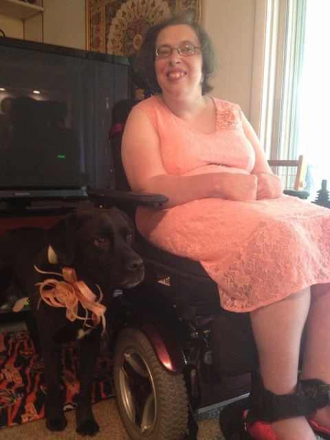 Susan Koller smiles from her wheelchair wearing a pink lace dress while her support dog Chia stands next to her wearing a matching floral collar