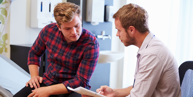 A young doctor talks to a young man sitting on an exam table.