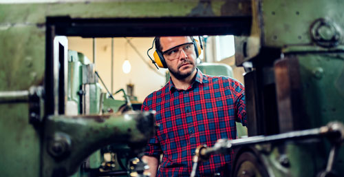 A white man stands at a machine in a factory wearing protective goggles and earmuffs 