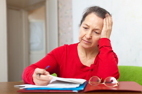 A white woman wearing a red sweater and holding a pen leans her face on her hand as she looks at a pile of papers.