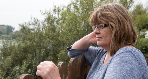 An older white woman with shoulder-length brown hair and glasses stands in front of a bush and looks over a wooden fence