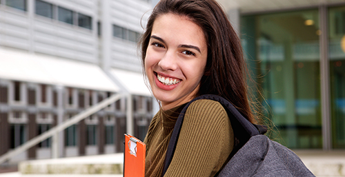 A white woman with long brown hair looks at the camera while holding an orange folder and carrying a gray backpack in front of a university building