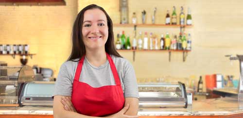 A woman with long brown hair and a red apron smiles as she stands in front of a counter at a cafe