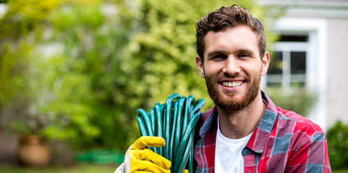 A young white man with brown hair and a beard smiles at the camera while wearing gardening gloves and holding a hose.